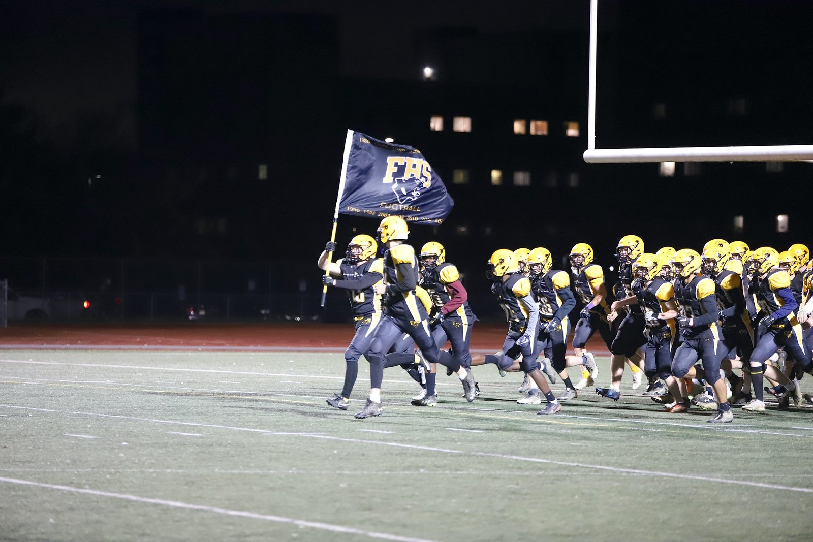The Black Kats run onto the field at night carrying a flag printed with the team's championship years
